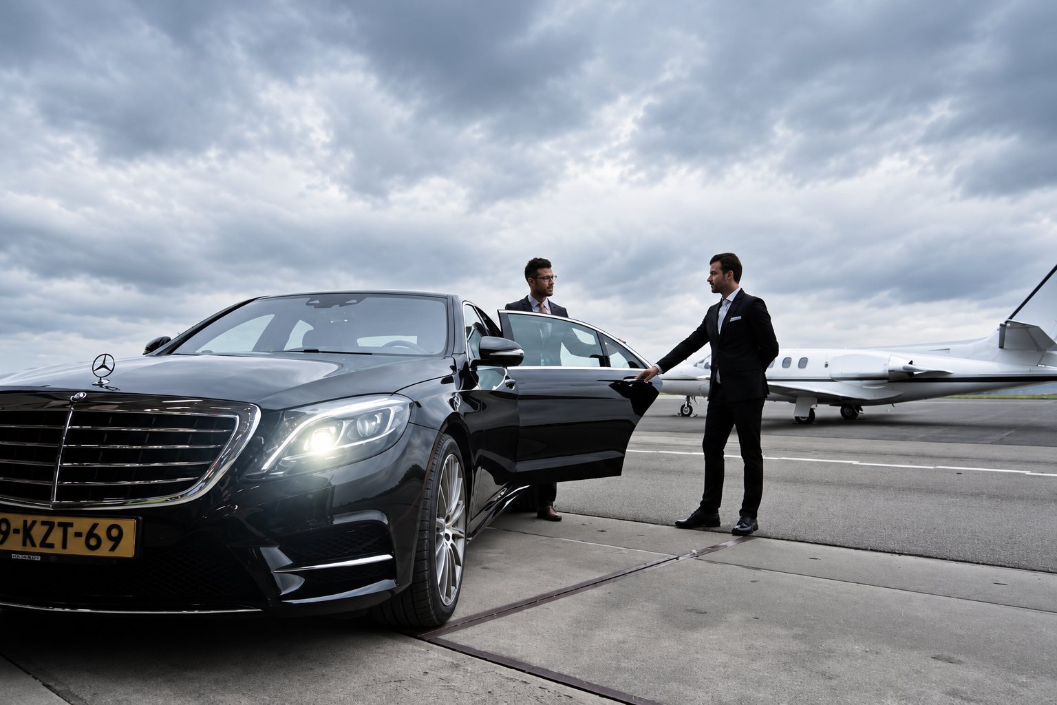 Sur une piste d’aéroport sous un ciel nuageux, un chauffeur en costume ouvre la porte d’une berline noire Mercedes, devant un jet privé et un autre homme en costume.