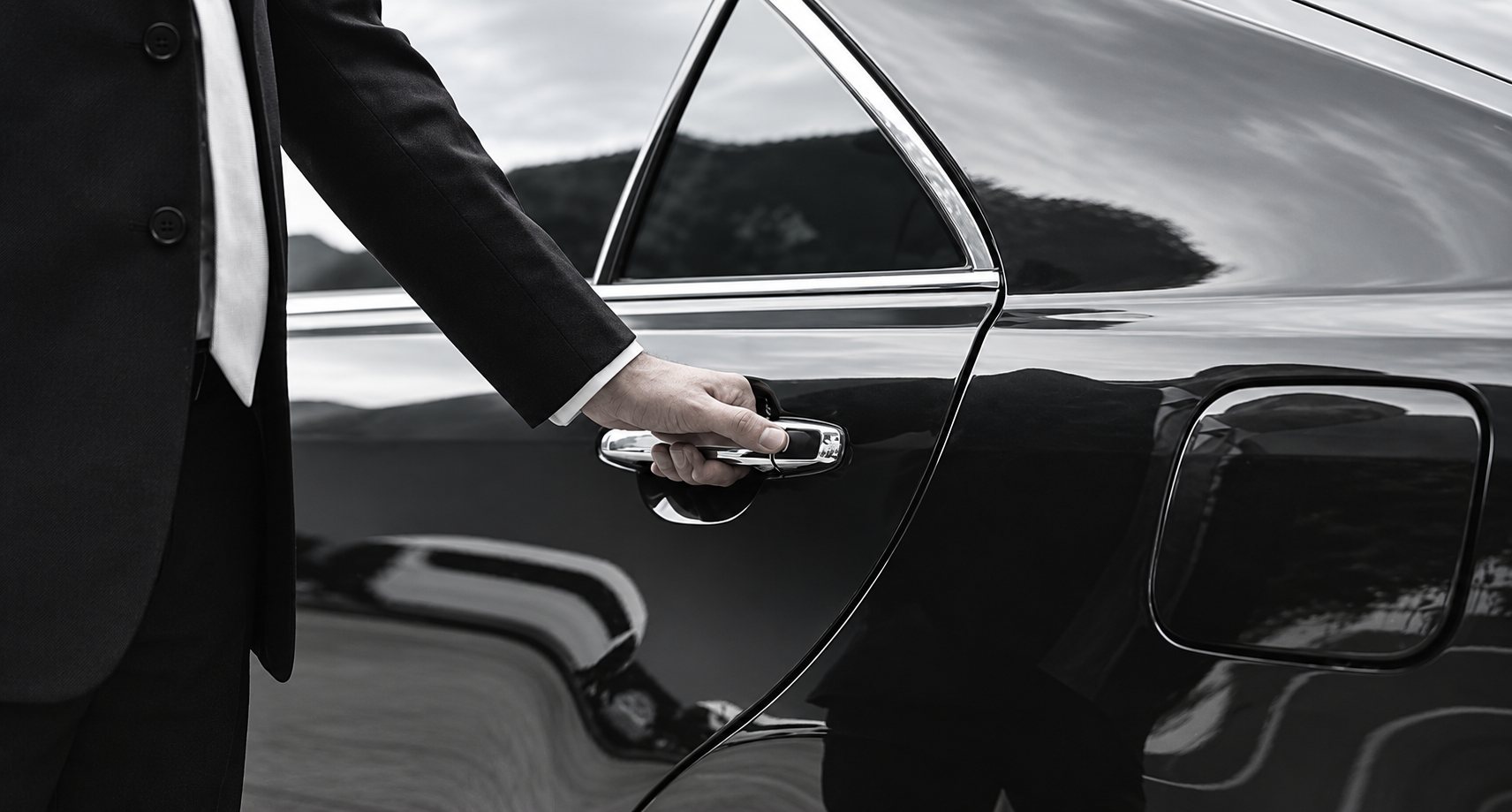 Un chauffeur en costume sombre ouvre la portière arrière d’une berline noire brillante, en noir et blanc.
